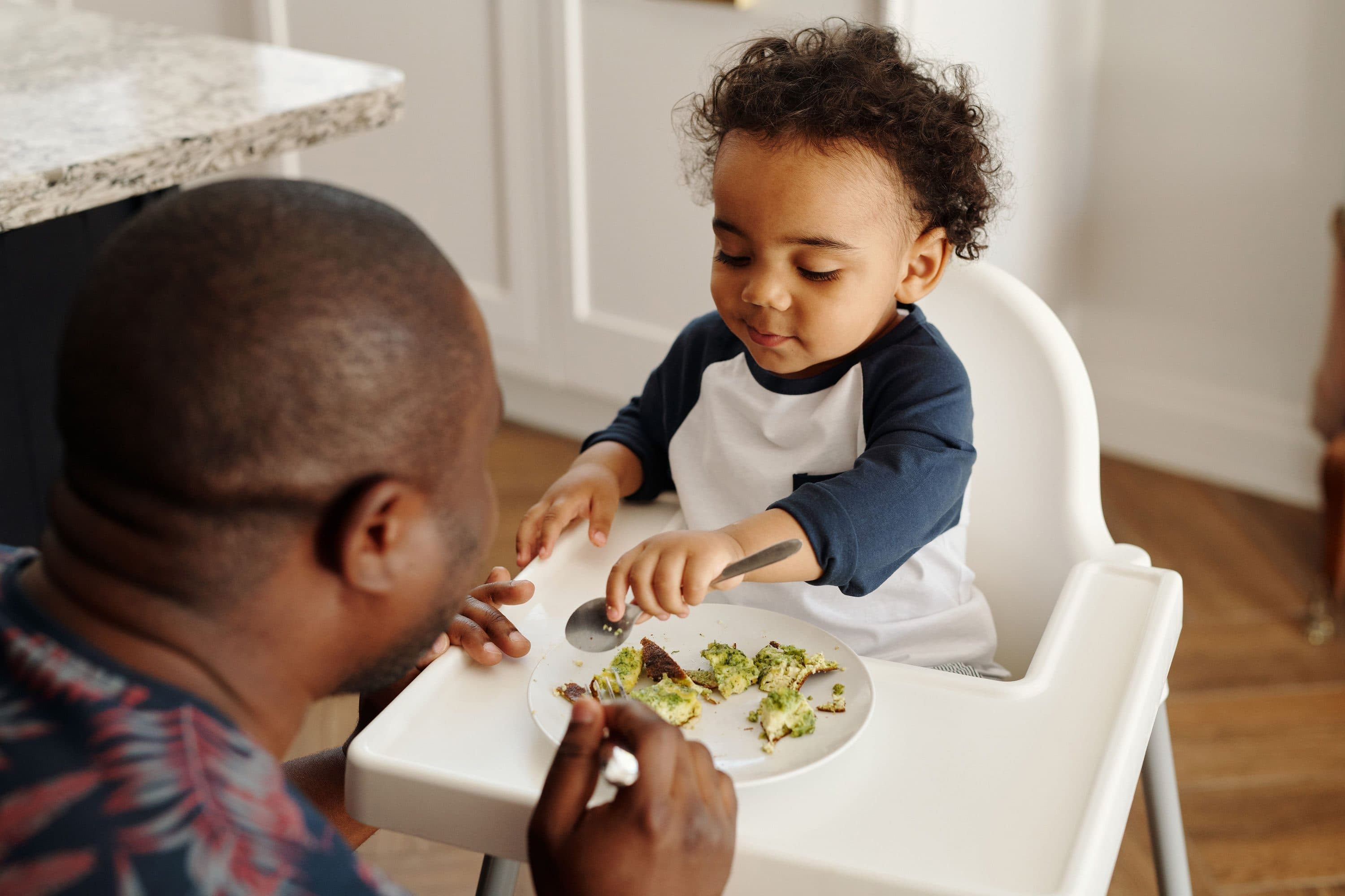 A father feeding his toddler, who is picking at a plate of greens and egg in a high chair