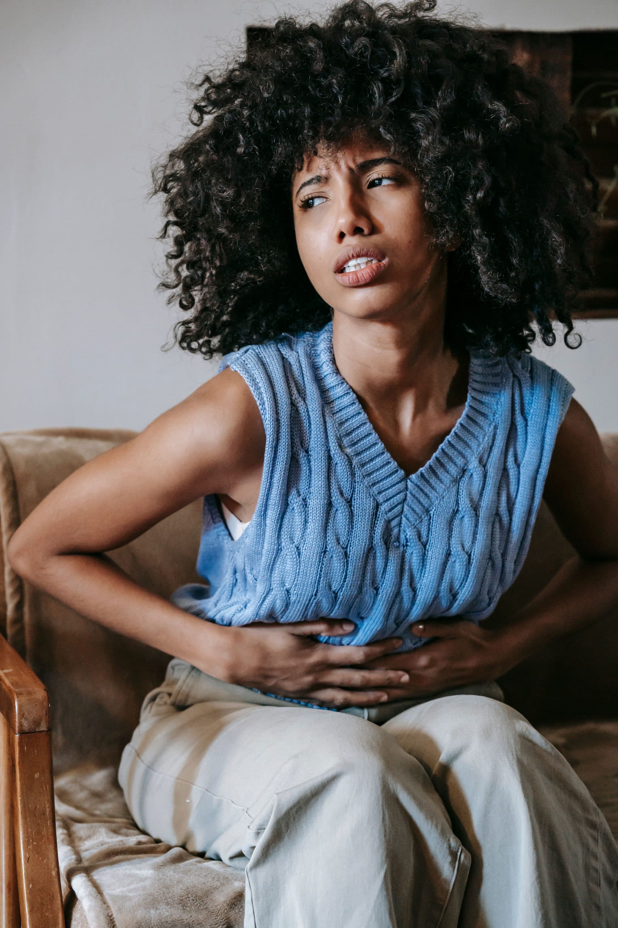A young woman sitting in an armchair with both hands on her lower abdomen, in discomfort