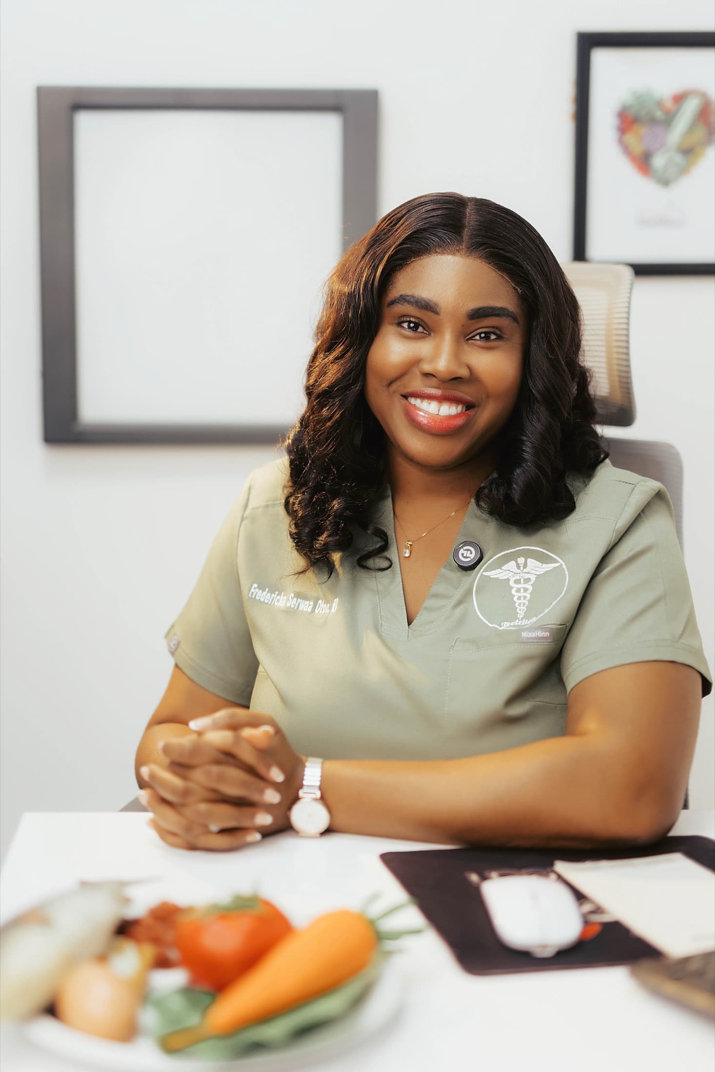 Fredericka Doku at her clinic desk in olive scrubs, a plate of vegetables in front of her